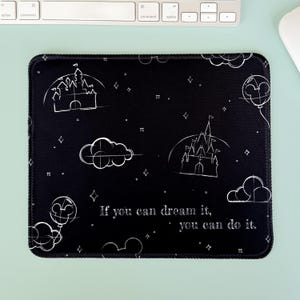 May include: Black mousepad with white line art of castles, clouds, stars, and Mickey Mouse balloons. The text "If you can dream it, you can do it" is printed on the pad. The mousepad has a black stitched border.