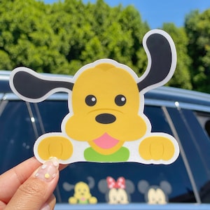 May include: A yellow cartoon dog sticker with black ears and a pink tongue. The sticker is on a car window.