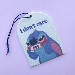 Könnte beinhalten: Ein weißes ovalförmiges Etikett mit einer blauen Schnur. Das Etikett zeigt ein Cartoon-Bild von Stitch aus Lilo & Stitch mit dem Text "i don't care." in lila.