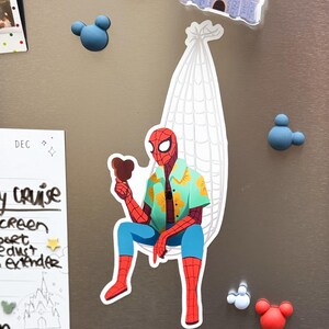 May include: A Spider-Man sticker wearing a Hawaiian shirt and blue pants, holding a chocolate ice cream bar, seated in a white web-like chair. Surrounding the sticker are Mickey Mouse-shaped magnets and a Disney castle magnet.