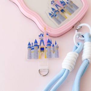 Op de afbeelding: Een lichtblauwe en witte telefoonband met een transparante bedel met een roze en blauw kasteel. De bedel is bevestigd aan een zilveren ring.