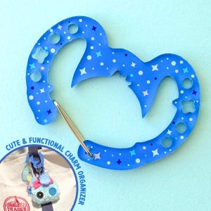 May include: A blue charm organizer with a star and dot pattern. The organizer has a unique shape with a metal clasp and the text "CUTE & FUNCTIONAL CHARM ORGANIZER". The organizer is shown with various charms attached.