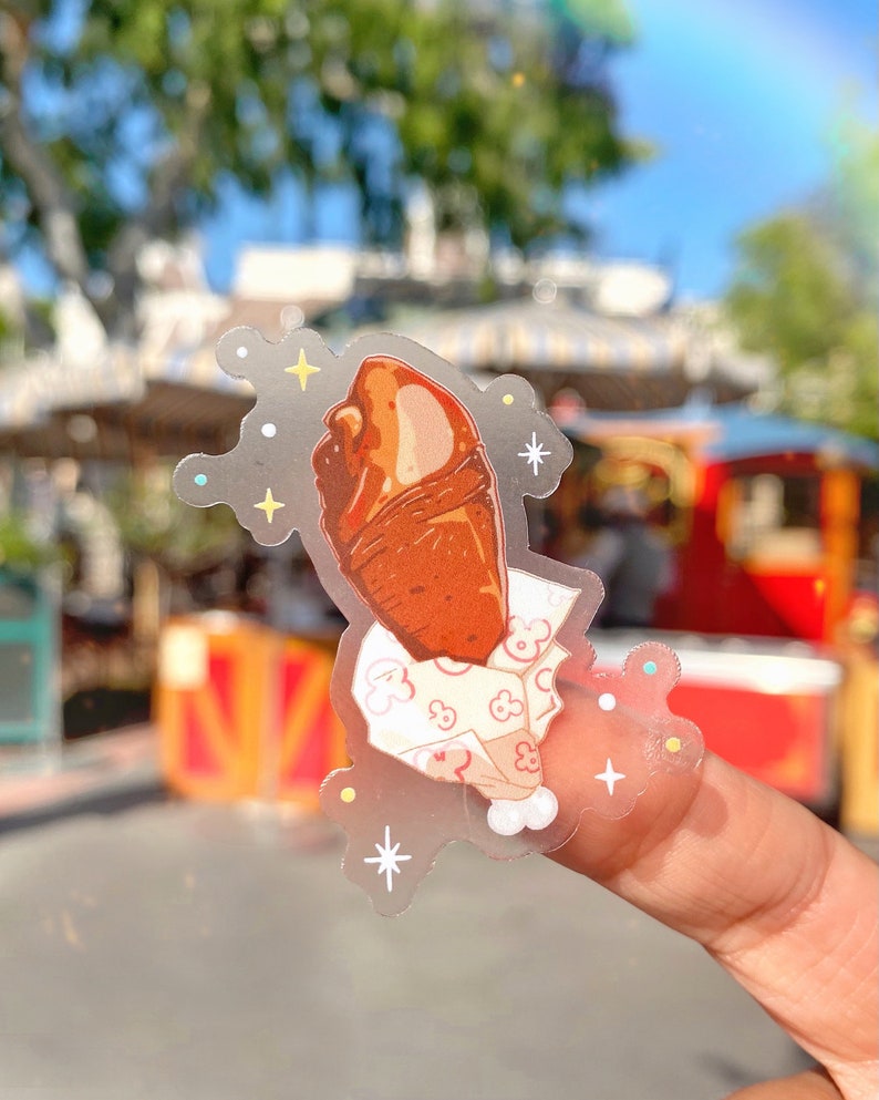 May include: A clear sticker featuring a cartoon illustration of a turkey leg. The turkey leg is brown with a white and pink paper wrap. The sticker has a decorative border with stars and sparkles.