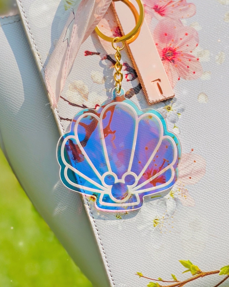 May include: A iridescent seashell keychain with a Mickey Mouse silhouette inside. The keychain is attached to a gold chain and a gold key ring.