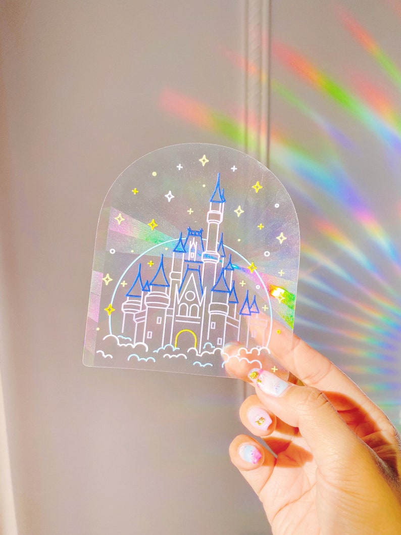 May include: A clear sticker with a holographic finish featuring a line drawing of Cinderella's castle with stars and clouds. The sticker is held in a hand with colorful nail polish.