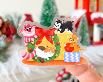 May include: A clear sticker featuring cartoon cats in a Christmas theme. The sticker shows a pink and white striped Cheshire Cat, a black cat, and two orange and white cats. A red bow and wreath surround a yellow gift box.