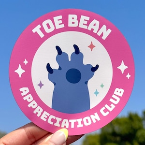 以下が含まれることがあります： ピンク色の縁と白い中心を持つ丸いステッカー。「TOE BEAN APPRECIATION CLUB」の文字が縁に印刷されています。青い足跡が中央にあり、4つの黒い爪があります。星のアクセントが含まれています。