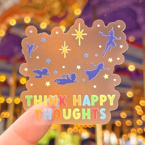 Op de afbeelding: Een transparante sticker met een gouden achtergrond en een regenboogkleurige tekst die "Think Happy Thoughts" zegt. De sticker bevat silhouetten van Peter Pan en Tinkerbell die met sterren vliegen.