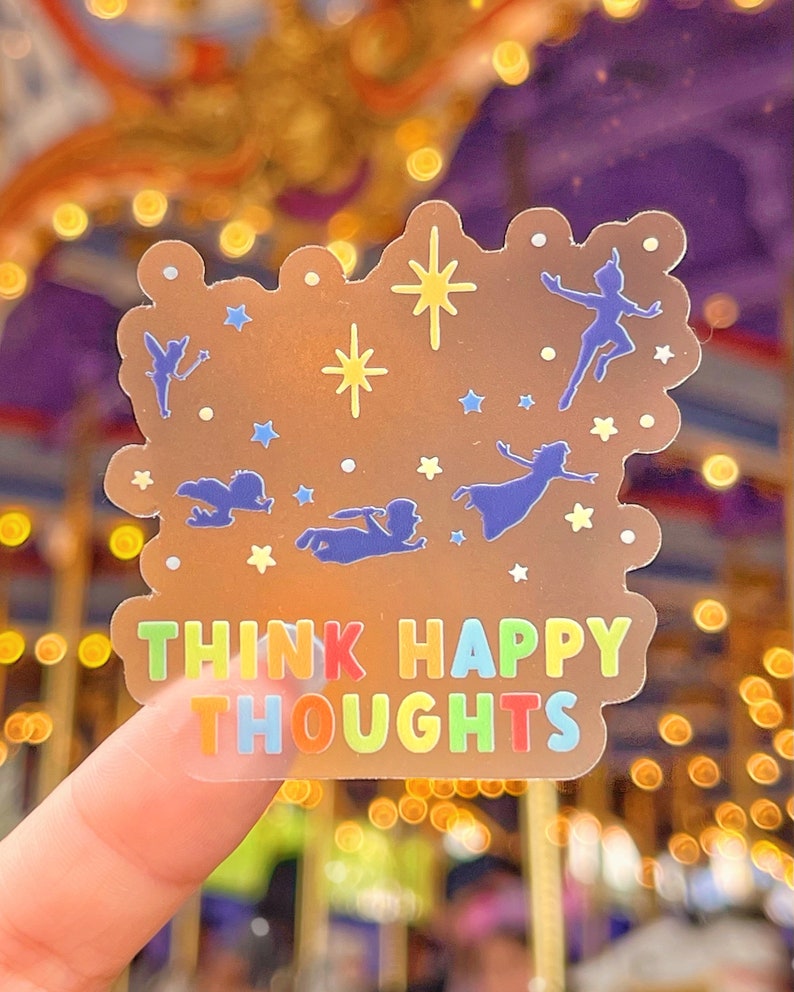 Op de afbeelding: Een transparante sticker met een gouden achtergrond en een regenboogkleurige tekst die "Think Happy Thoughts" zegt. De sticker bevat silhouetten van Peter Pan en Tinkerbell die met sterren vliegen.