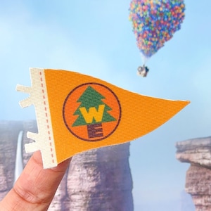 Puede incluir: Bandera de banderín naranja con un emblema circular que presenta un árbol verde y la letra "W". La bandera tiene un borde blanco con una línea punteada. En el fondo, un grupo de globos coloridos flota en el cielo.