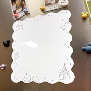 以下が含まれることがあります： お城と雲のデザインが施された、白い波型の縁取りのメモボード。ボードは小さな星とひし形の形で装飾されています。ミッキーマウスをテーマにしたマグネットと写真が金属の表面に付いています。