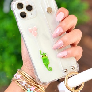 May include: A clear phone case with a green chameleon design and pink Mickey Mouse balloons. The case has a white strap with a gold ring. The phone case is being held by a hand with pink and white nail polish.