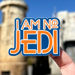 May include: A blue and orange sticker with the text "I AM NO JEDI". The text is outlined in white. The sticker is held up against a blurred background of buildings and a blue sky.