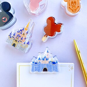 May include: A selection of clear acrylic Disney-themed accessories, including a Mickey Mouse head, a pink Minnie Mouse bow, a castle, and a Mickey-shaped ice cream bar. A gold pen and a notepad with a castle clip are also present.