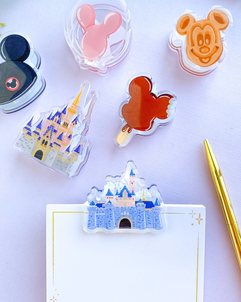May include: A selection of clear acrylic Disney-themed accessories, including a Mickey Mouse head, a pink Minnie Mouse bow, a castle, and a Mickey-shaped ice cream bar. A gold pen and a notepad with a castle clip are also present.