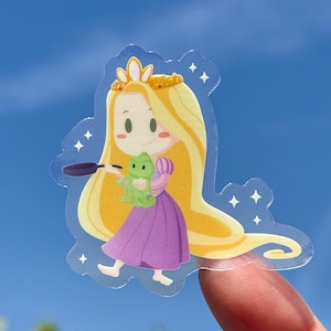 Op de afbeelding: Een transparante sticker met een cartoonillustratie van een prinses met lang blond haar, die een paarse jurk en een gouden kroon draagt. Ze houdt een koekenpan in de ene hand en een groene kikker in de andere. De sticker heeft een glinsterende achtergrond.