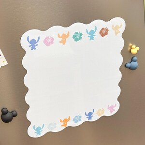 May include: White, scalloped-edge memo board with colorful Stitch and flower designs. The board has a blank center for notes and is decorated with blue, pink, orange, and brown graphics. Magnetic Mickey Mouse shapes are on the right and left.