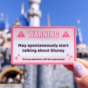 May include: Pink warning sticker with a white exclamation point and the text "WARNING! May spontaneously start talking about Disney. Strong opinions will be expressed." The sticker is held in front of a blurred image of a blue and white castle.