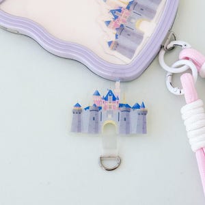 Op de afbeelding: Een transparante plastic ID-badgehouder met een roze en blauwe kasteel illustratie. De houder heeft een zilveren D-ring en een roze en witte lanyard.