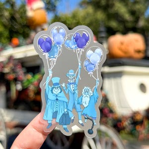 May include: A clear sticker featuring three cartoon ghosts holding blue and purple Mickey Mouse-shaped balloons. The ghosts are dressed in light blue attire. The sticker is held up by a hand.