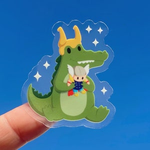 May include: A green crocodile with yellow horns and a white and red superhero doll in its arms. The crocodile is holding the doll in front of its chest. The background is a blue sky with white stars.