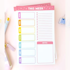 Könnte beinhalten: Ein rosa Wochenplaner mit bunten Wochentagen und einem Notizbereich. Der Planer hat einen rosa Hintergrund mit weißen Sternen und den Text "THIS WEEK" in Schwarz.