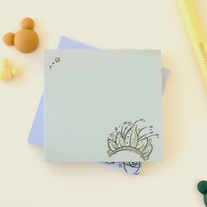 May include: A light blue square sticky note with a floral design in the corner. The design is outlined in black and filled with tan and gold. A yellow pen and two small Mickey Mouse-shaped erasers are in the background.
