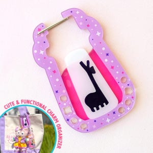 Könnte beinhalten: Ein lila Charm-Organizer mit einem silbernen Verschluss, verziert mit Stern- und Punktmustern. Er verfügt über einen weißen und rosa Einsatz mit einer schwarzen Lama-Silhouette. Der Text "CUTE & FUNCTIONAL CHARM ORGANIZER" ist sichtbar.