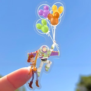 Op de afbeelding: Een transparante sticker met Woody en Buzz Lightyear uit Toy Story, omhooggehouden door Mickey Mouse-vormige ballonnen in paars, groen en oranje. De personages zweven in de lucht tegen een blauwe lucht.