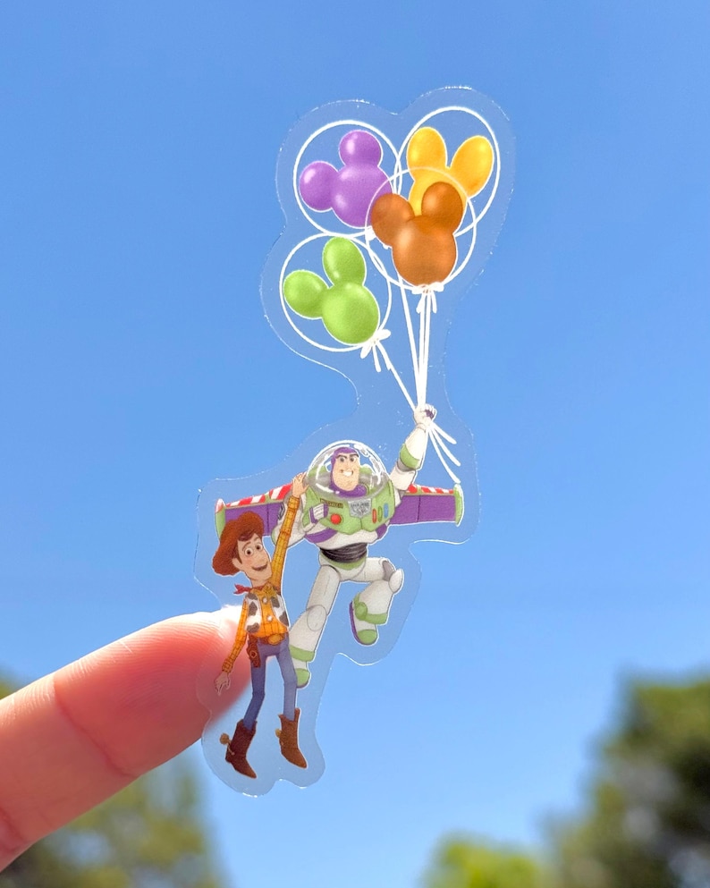 Op de afbeelding: Een transparante sticker met Woody en Buzz Lightyear uit Toy Story, omhooggehouden door Mickey Mouse-vormige ballonnen in paars, groen en oranje. De personages zweven in de lucht tegen een blauwe lucht.