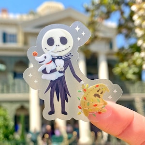 May include: A clear sticker featuring Jack Skellington from The Nightmare Before Christmas holding Zero, his ghost dog. The sticker also features a small, round, brown character with Christmas lights wrapped around it.