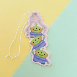 Op de afbeelding: Een grillige luchtverfrisser met drie groene aliens in blauwe outfits, hangend aan een wit touwtje. De aliens zijn gestapeld, met een grijpmachine-afbeelding bovenaan. De achtergrond is verdeeld in geel en lichtgroen.