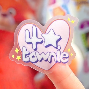 Può includere: Un adesivo trasparente a forma di cuore con sfondo rosa. L'adesivo presenta una stella bianca, il numero "4" e la parola "townie" in un carattere viola in 3D. L'adesivo ha una finitura scintillante e iridescente.