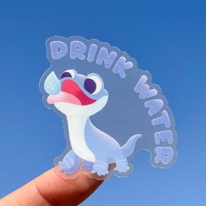 May include: A clear sticker featuring a blue cartoon lizard drinking water. The sticker reads "Drink Water" in a purple font.