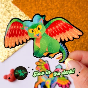 Peut inclure: Un autocollant vert et rouge qui brille dans le noir repr&eacute;sentant une cr&eacute;ature mythique ail&eacute;e. L'autocollant a un contour noir et est tenu par une main. Le texte "Glow in the dark!" est visible sur l'image.