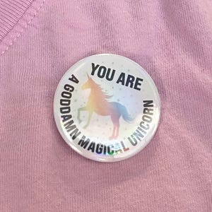 May include: A circular badge with the words "YOU ARE A GODDAMN MAGICAL UNICORN" around a colourful unicorn image. The badge has a holographic finish and is set against a pink backdrop.