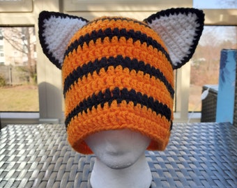 May include: A crocheted tiger hat with orange and black stripes and white ears. The hat is on a white mannequin head.