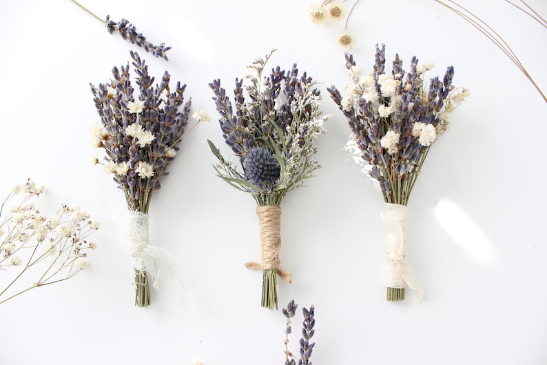 May include: Three small bouquets of dried lavender and white flowers tied with twine and ribbon. The bouquets are arranged in a row on a white surface.