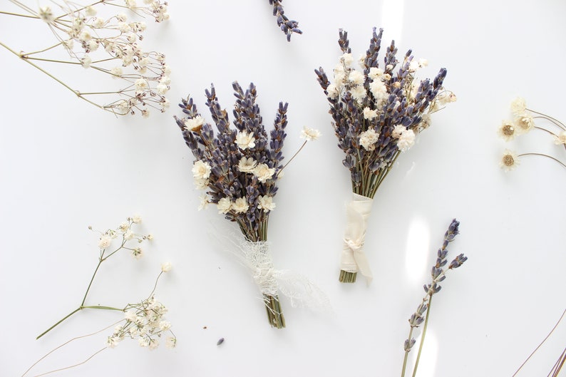 May include: Two small bouquets of dried lavender and white flowers tied with twine and ribbon. The bouquets are on a white background.