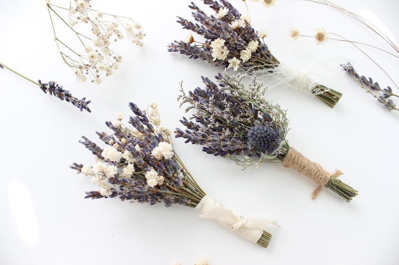 May include: Three bouquets of dried lavender and white flowers tied with twine and ribbon. The bouquets are arranged on a white surface.