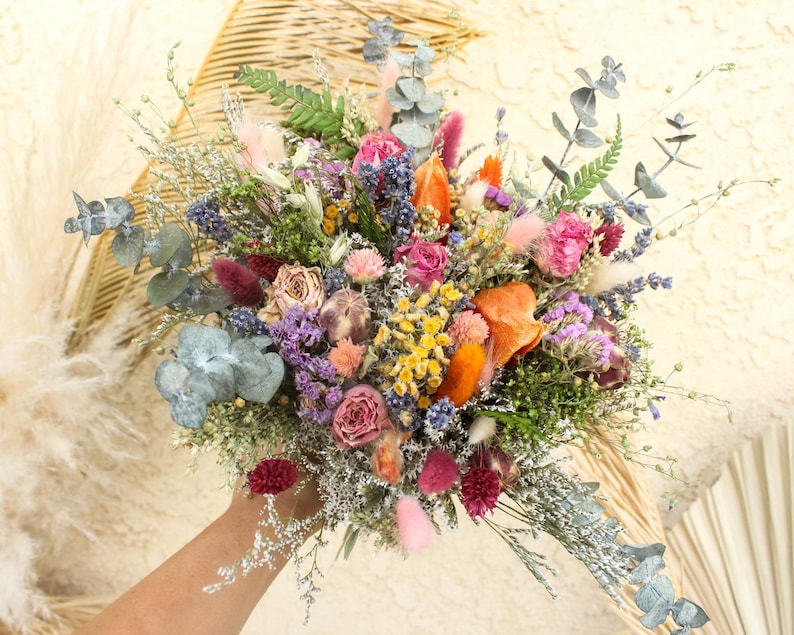 May include: A vibrant bouquet of dried flowers in shades of pink, orange, purple, and yellow. The arrangement includes roses, eucalyptus, and other greenery, held in a hand. The background is a neutral cream color.