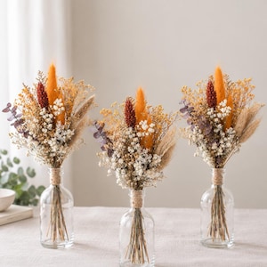 May include: Three clear glass bottles, each containing a dried floral arrangement featuring orange and rust-coloured plumes, white baby's breath, and eucalyptus leaves. The stems are tied with twine at the neck of each bottle.
