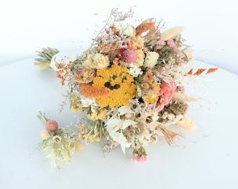 May include: A dried flower bouquet and boutonniere featuring a variety of textures and colors. The bouquet includes yellow, pink, and cream-colored flowers, with accents of orange and brown. The boutonniere mirrors the bouquet's design, creating a cohesive floral set.