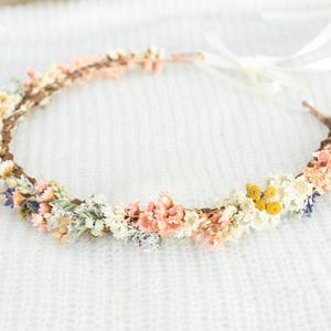 May include: A floral crown featuring a variety of dried flowers in shades of peach, yellow, white, and blue. The flowers are arranged on a brown base and tied with white ribbons, perfect for weddings or special occasions.