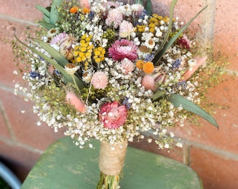 Puede incluir: Un colorido ramo de flores secas, que incluye flores rosas, amarillas y blancas, envuelto con hilo. El arreglo presenta una variedad de texturas y formas, creando una estética rústica y natural. El ramo está sobre una superficie verde.