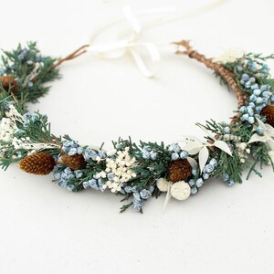 May include: A circular floral crown featuring green sprigs, blue berries, white accents, and small brown pinecones. The crown is tied with a white ribbon. This decorative headpiece is suitable for weddings or special occasions.
