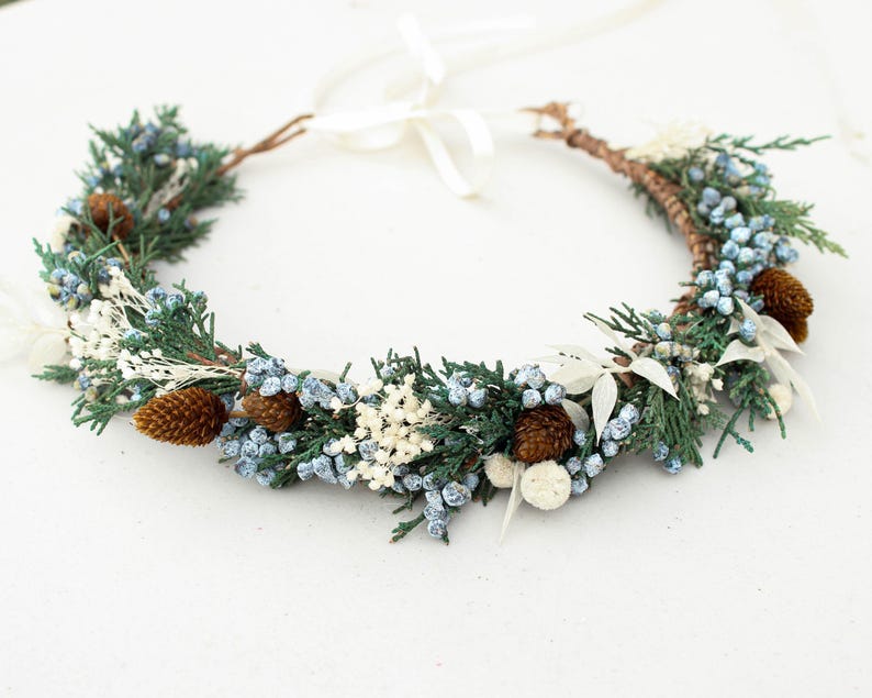 May include: A circular floral crown featuring green sprigs, blue berries, white accents, and small brown pinecones. The crown is tied with a white ribbon. This decorative headpiece is suitable for weddings or special occasions.