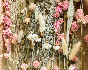 May include: A bouquet of dried flowers in shades of pink and white, including delicate blooms, fluffy seed heads, and long, slender stems. The flowers are arranged in a natural, rustic style, creating a charming and elegant display.