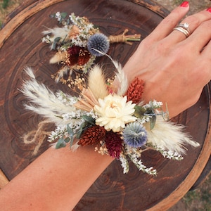 Könnte beinhalten: Ein Handgelenkscorsage aus getrockneten Blumen in Braun-, Weiß- und Burgunder-Tönen. Das Corsage enthält getrocknete Gräser, kleine weiße Blüten und eine große weiße Blüte in der Mitte.