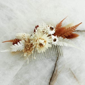 May include: A floral hair comb featuring a cluster of dried flowers and foliage. The arrangement includes cream-colored daisies, white ferns, and rust-colored accents. The comb is made of silver-toned metal and is designed for bridal or special occasion hairstyles.